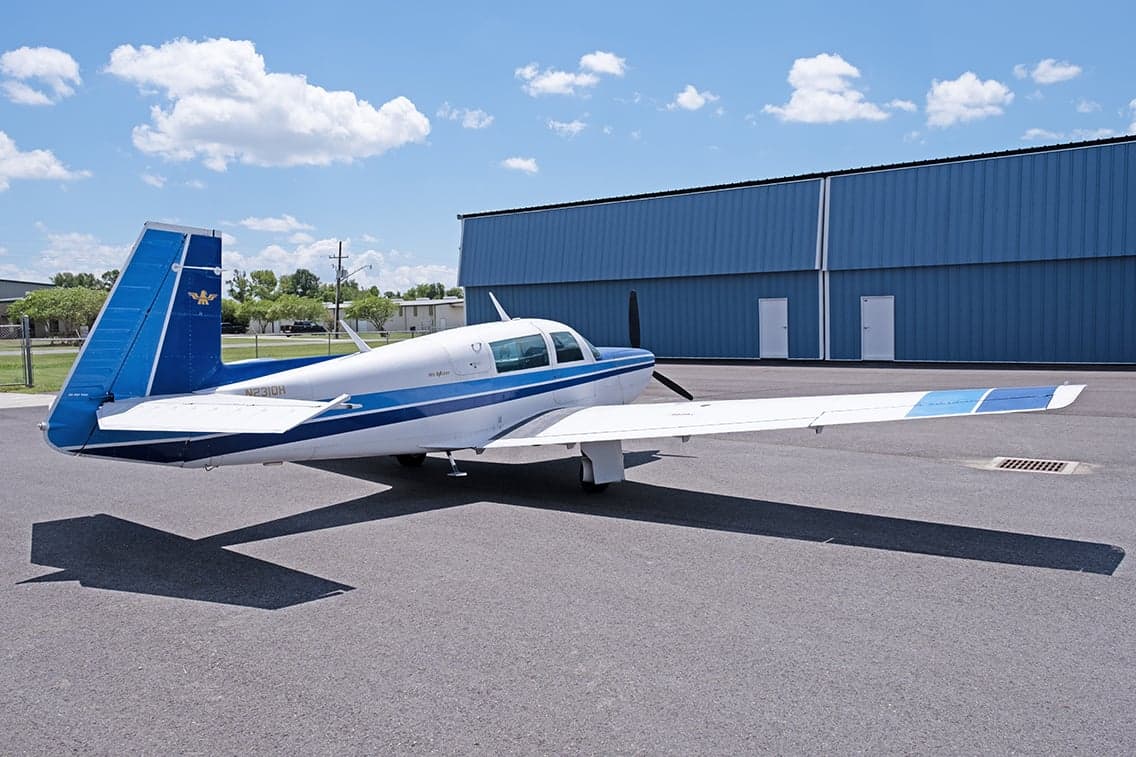Side view of a white and blue Mooney M20F aircraft parked on the tarmac in front of a hangar.
