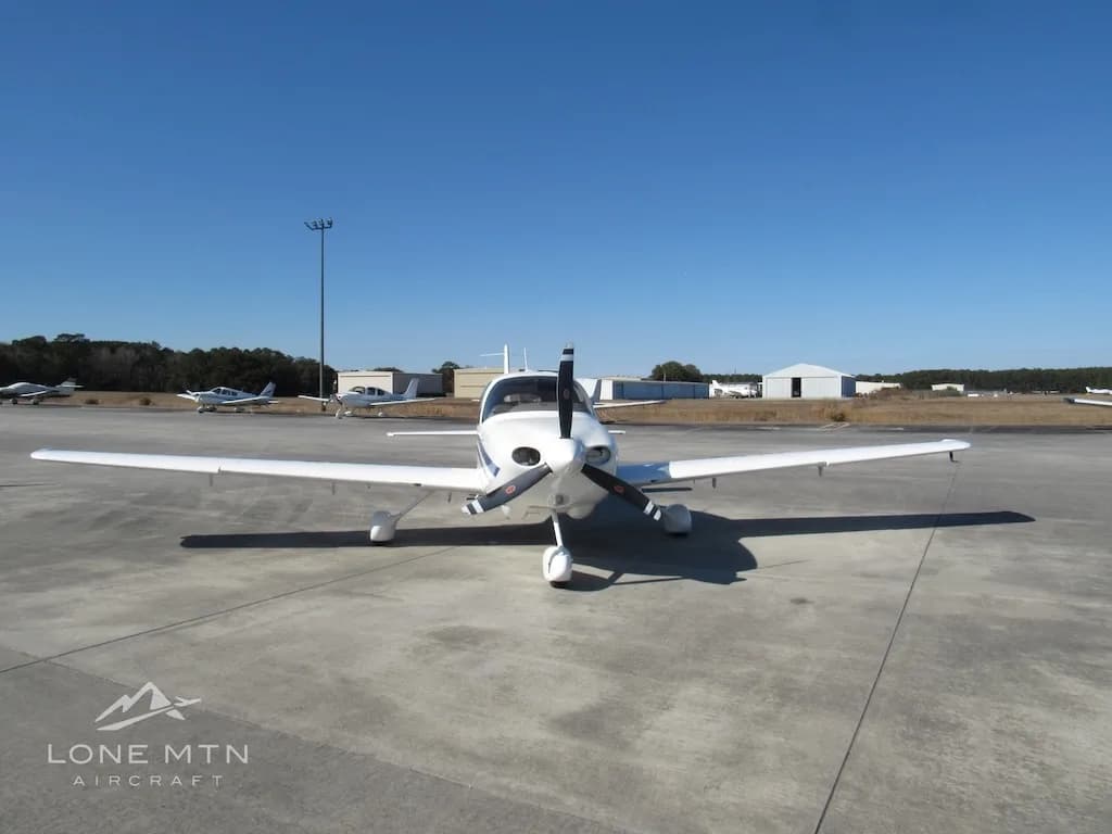 A head-on view of a white Cirrus SR20 aircraft with blue and grey stripes parked on a concrete ramp.