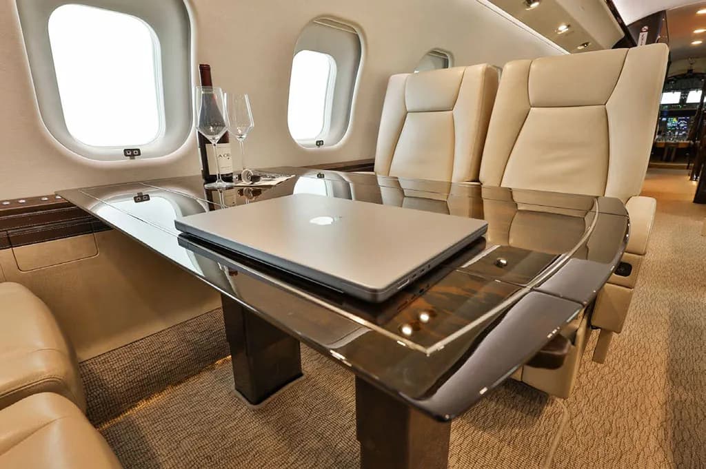 A close-up of a polished wooden table in the business jet cabin, with a laptop, wine bottle, and glasses.