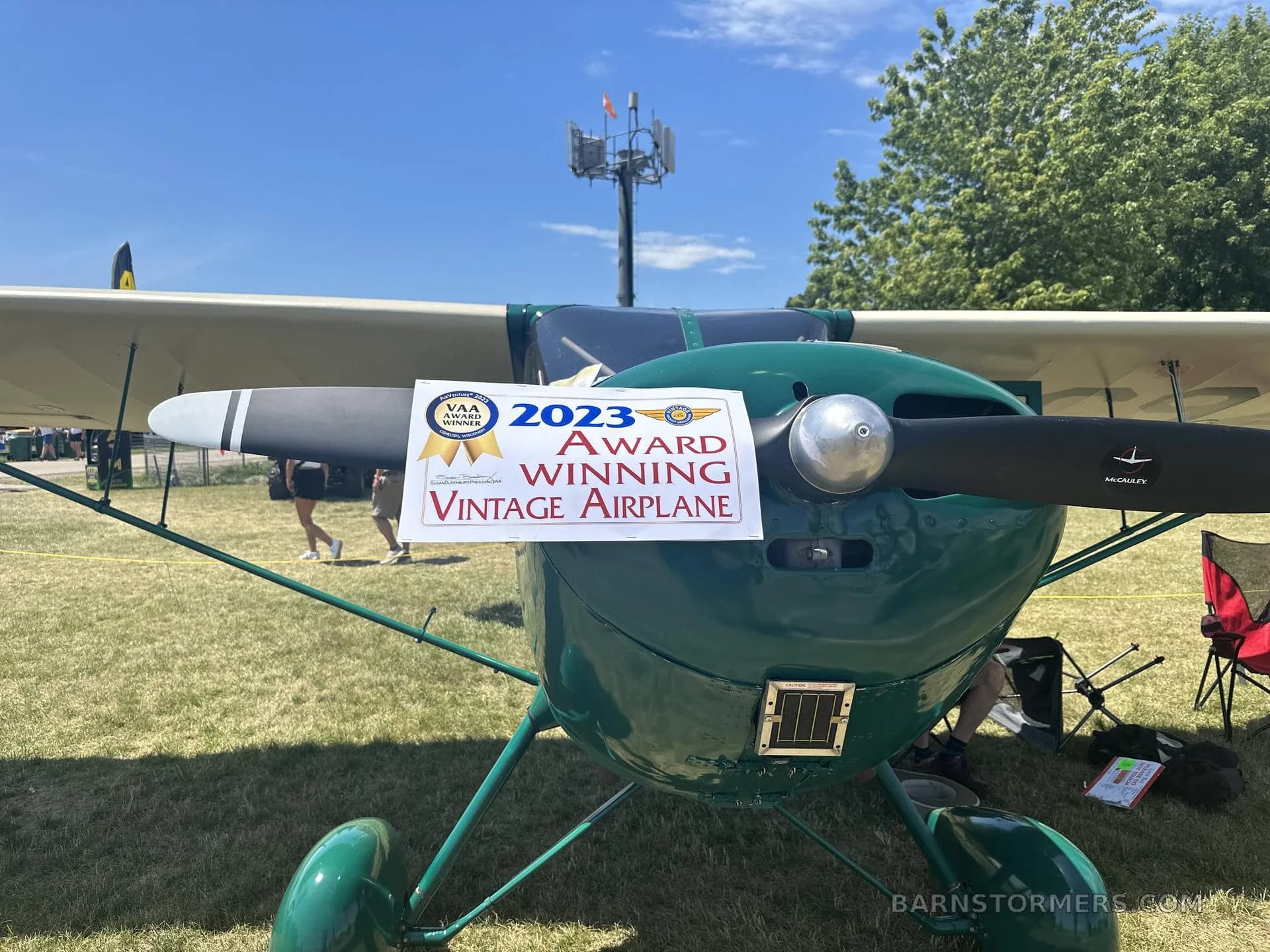 A close-up of the front of a green vintage aircraft, showing its propeller and an award sign for "2023 Award Winning Vintage Airplane".