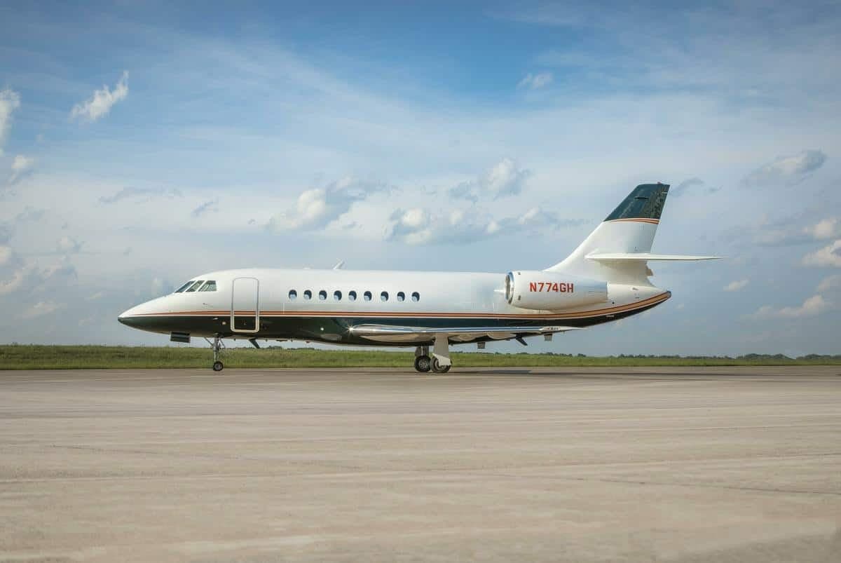 A white and dark green Dassault Falcon business jet (N774GH) with orange stripes on a tarmac under a partly cloudy sky.