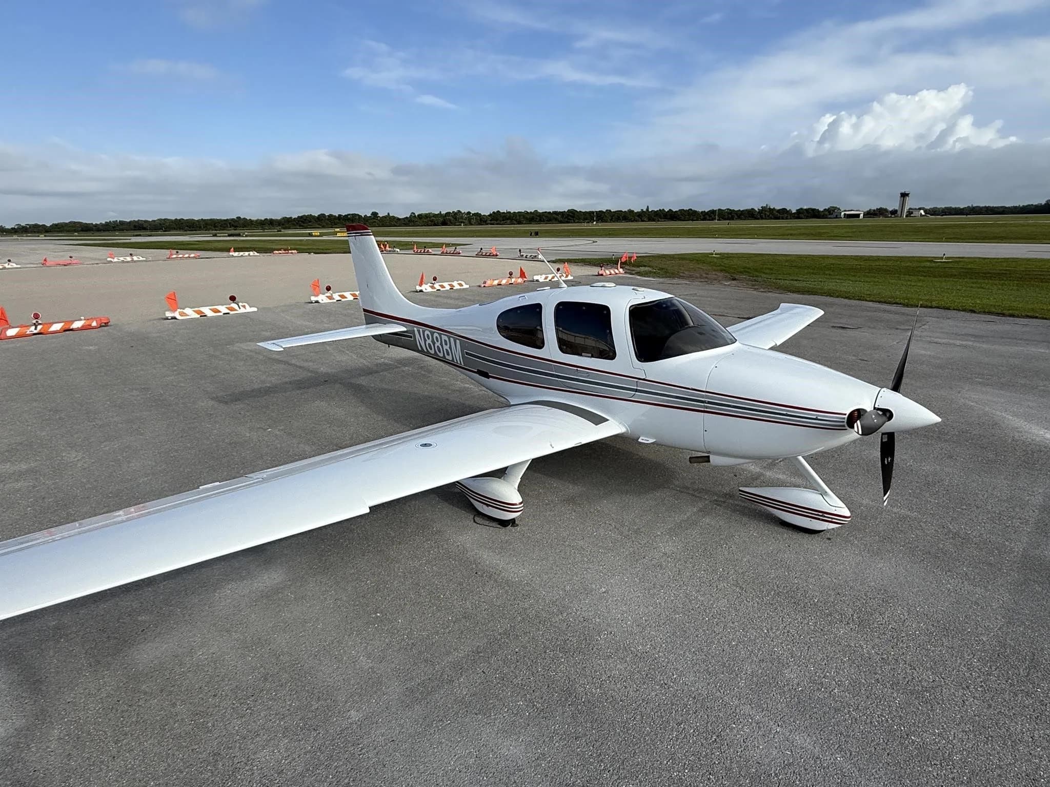 A white Cirrus SR22 aircraft with red and gray stripes, tail number N88BM, parked on an airport tarmac under a partly cloudy sky.