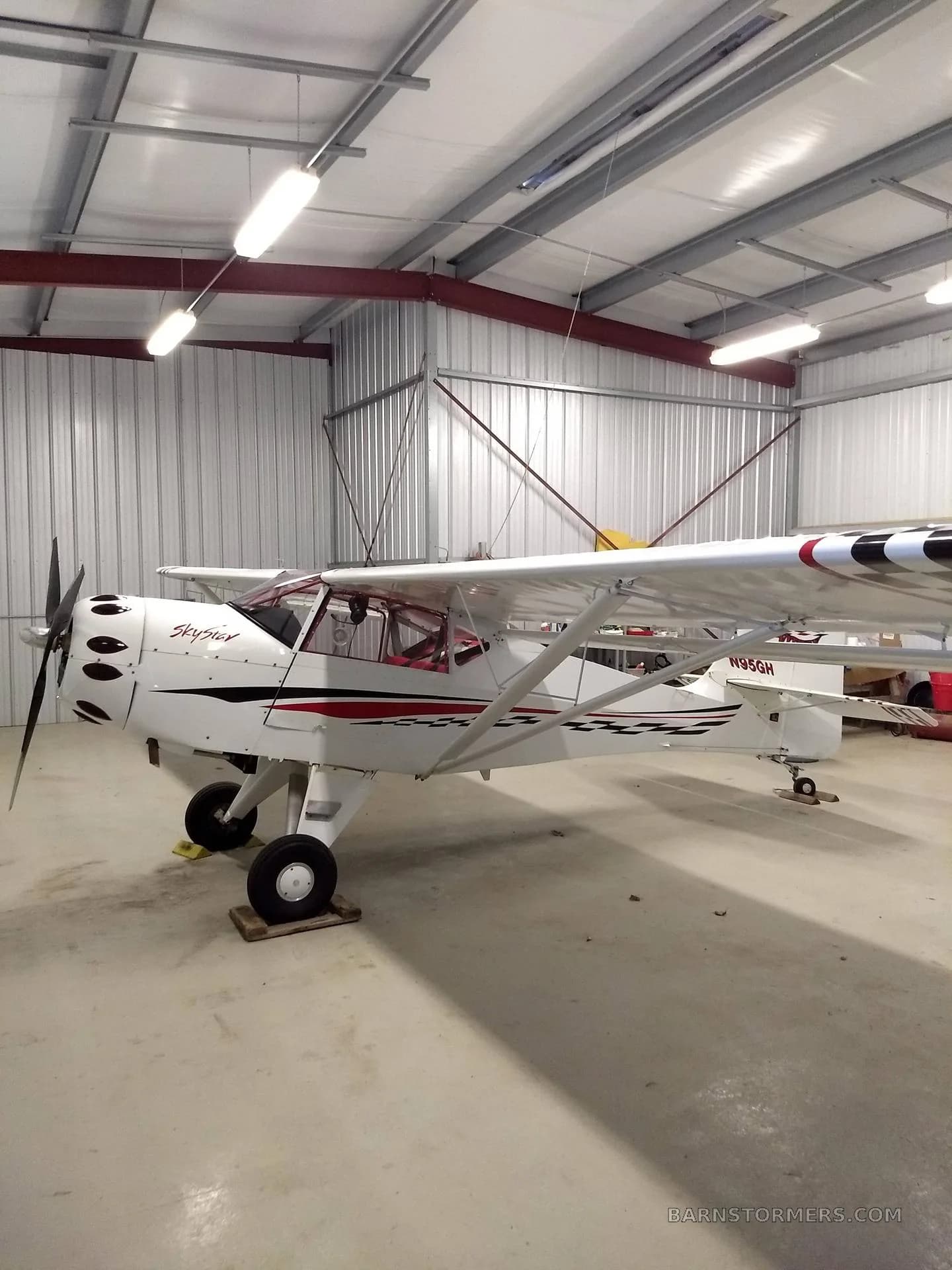 A white SkyStar Kitfox aircraft with red and black checkerboard accents is parked inside a hangar.