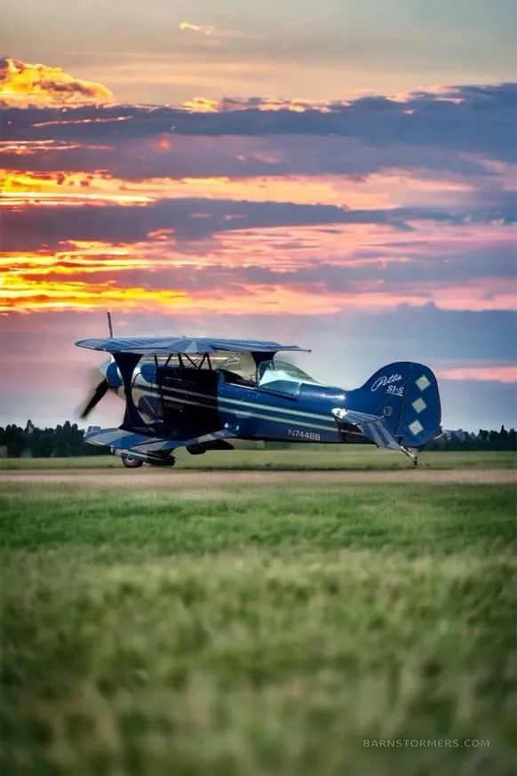 A blue Pitts S1-S biplane with white and green accents takes off from a grassy field at sunset.
