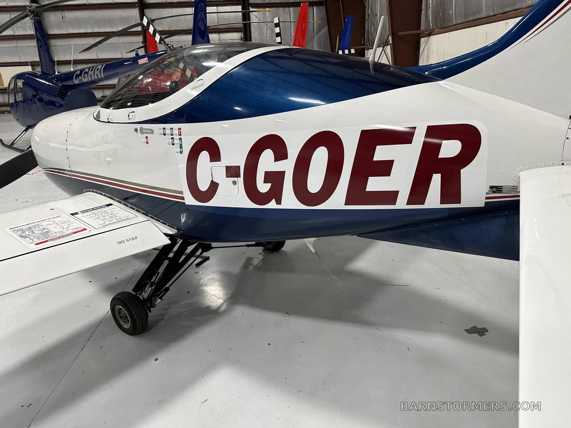 A close-up side view of a white and blue aircraft with registration C-GOER in red, parked inside a hangar next to a blue helicopter.