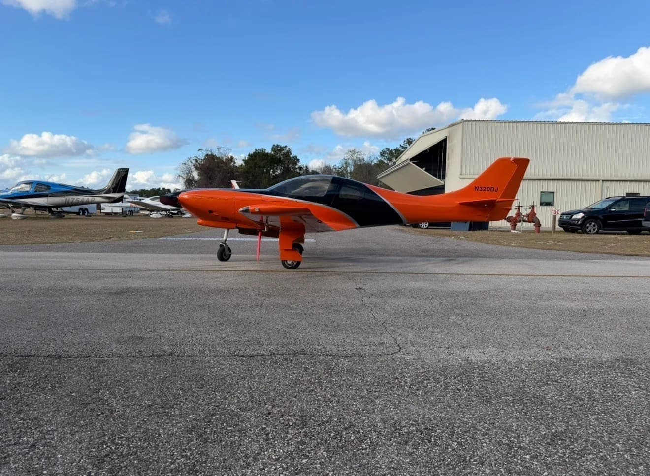 The orange and black experimental aircraft, N320DJ, is parked on an airfield tarmac with other planes and hangars in the background.