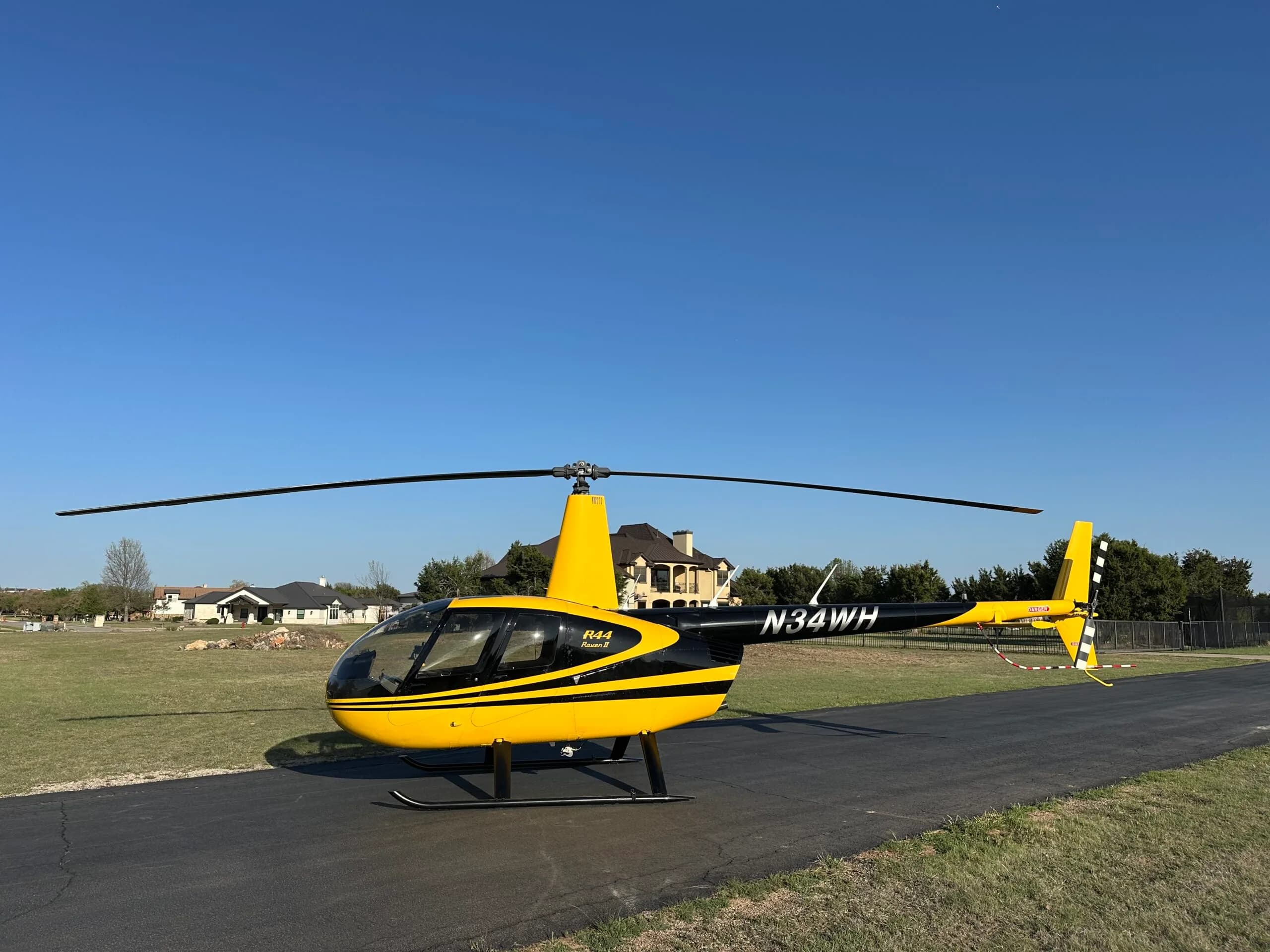 A yellow and black Robinson R44 Raven II helicopter with registration N34WH is parked on an asphalt strip under a clear blue sky.