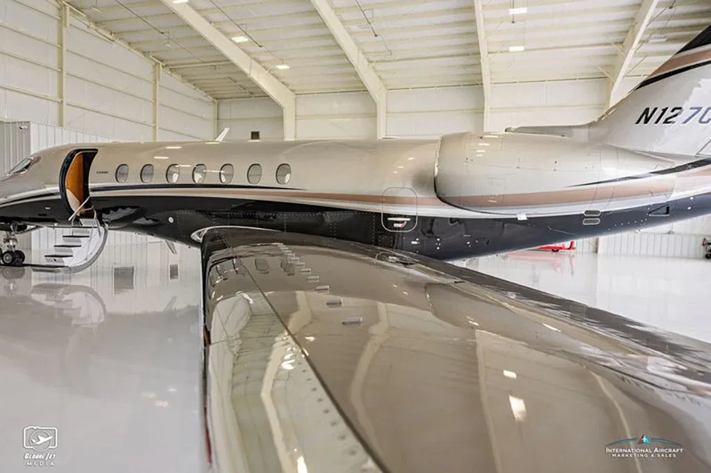 A close-up side view of the fuselage and wing of a grey and black business jet with its door open, inside a hangar.