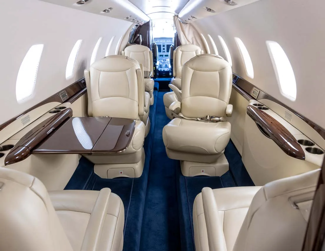 A view down the aisle of a business jet's cabin, showing cream leather seats, fold-out tables, and large windows.
