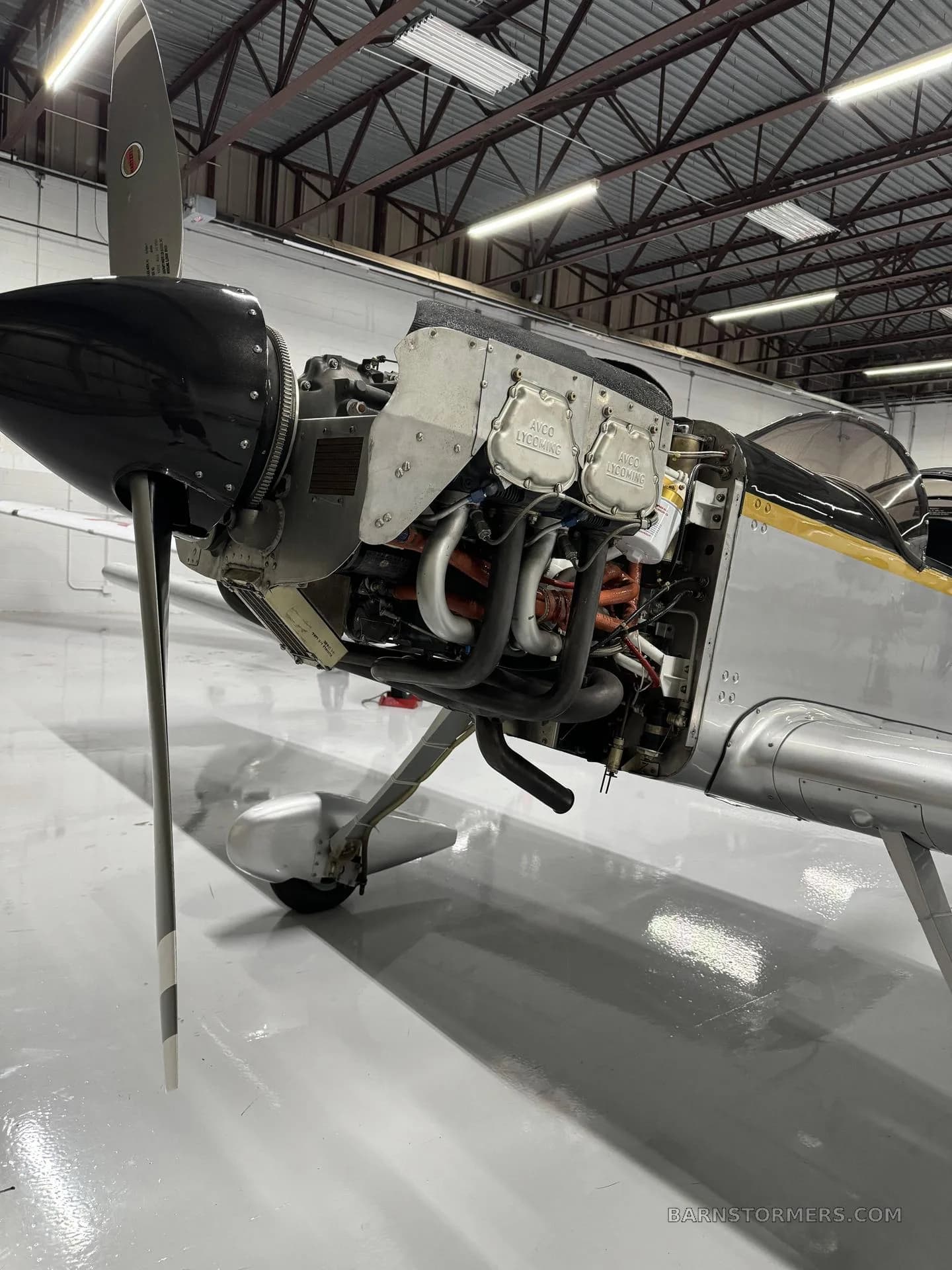 The engine of a silver aircraft is exposed with the cowling removed, showing the Lycoming engine and exhaust pipes.