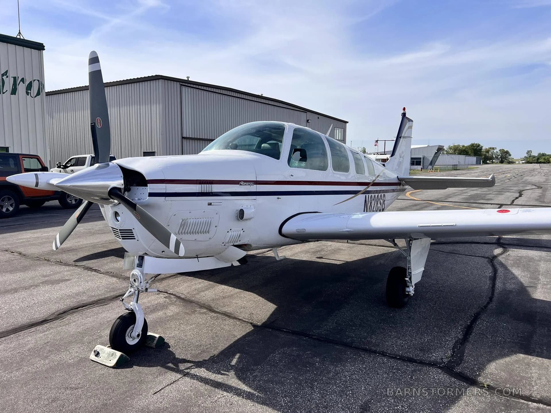 A white Beechcraft Bonanza with red and blue stripes is parked on a tarmac in front of a hangar.