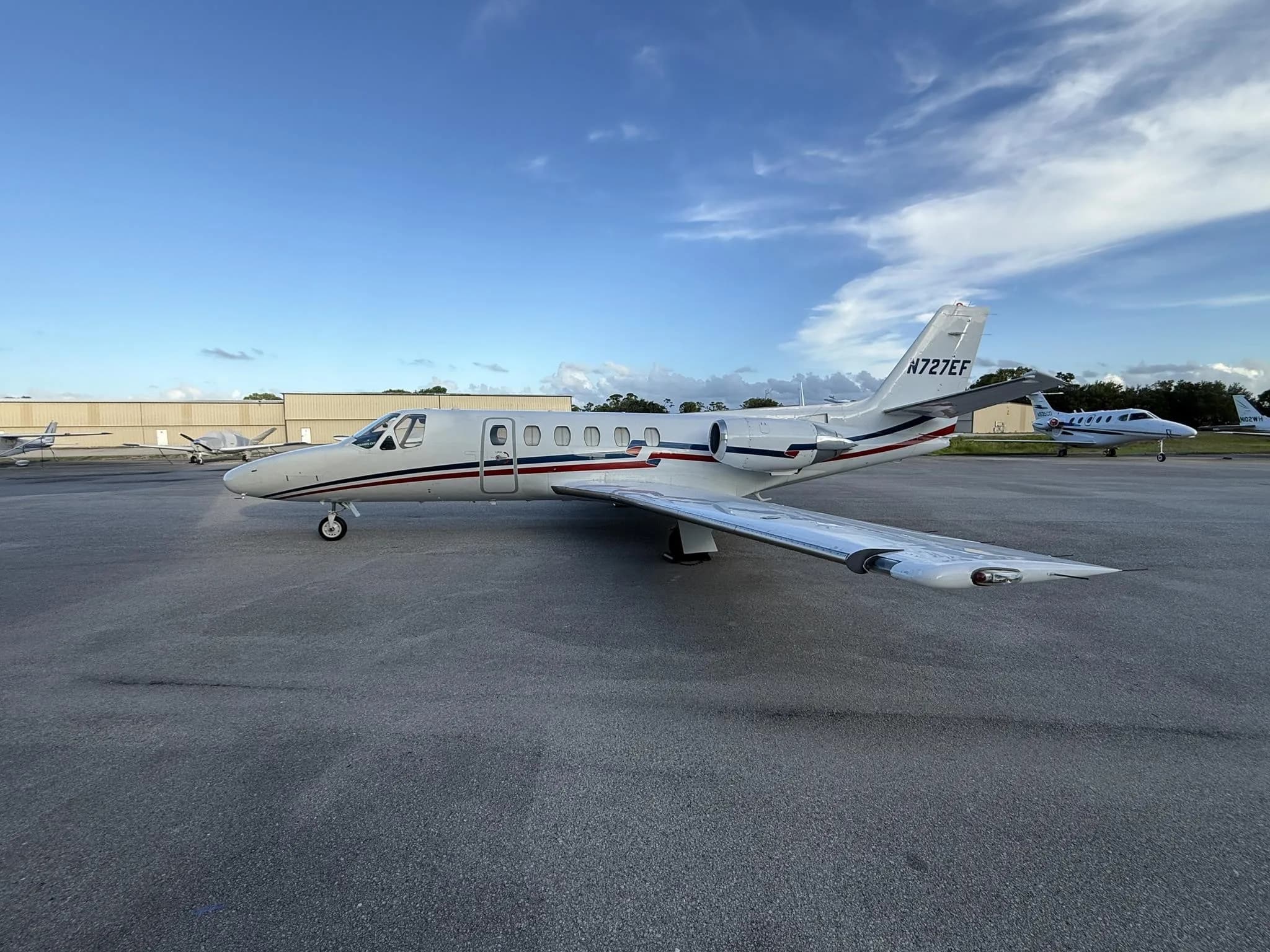 A white Cessna Citation business jet (N727EF) with red and blue stripes parked on an airfield under a blue sky.