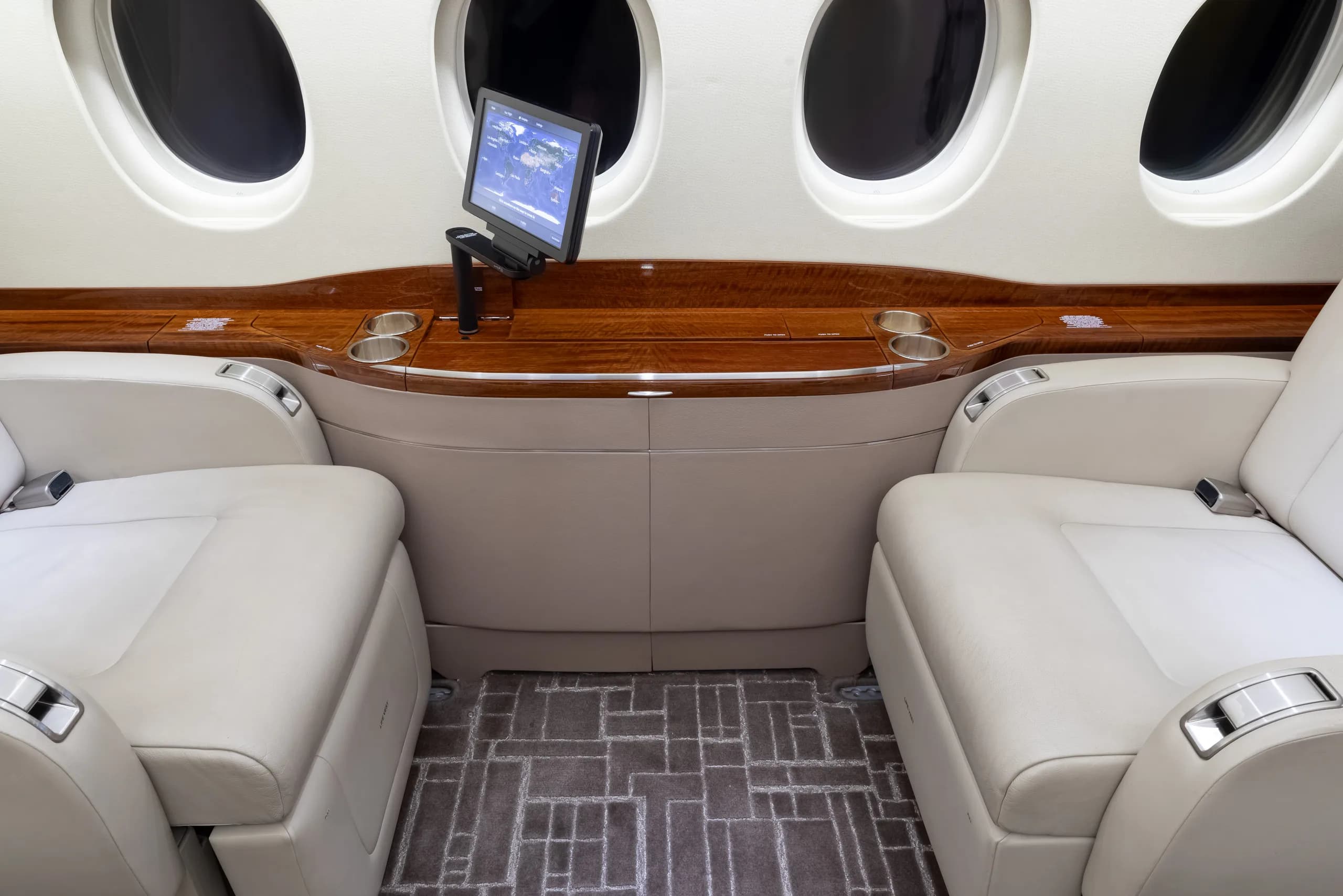 Close-up view of two light beige leather aircraft seats facing a polished wooden console with a retractable screen.