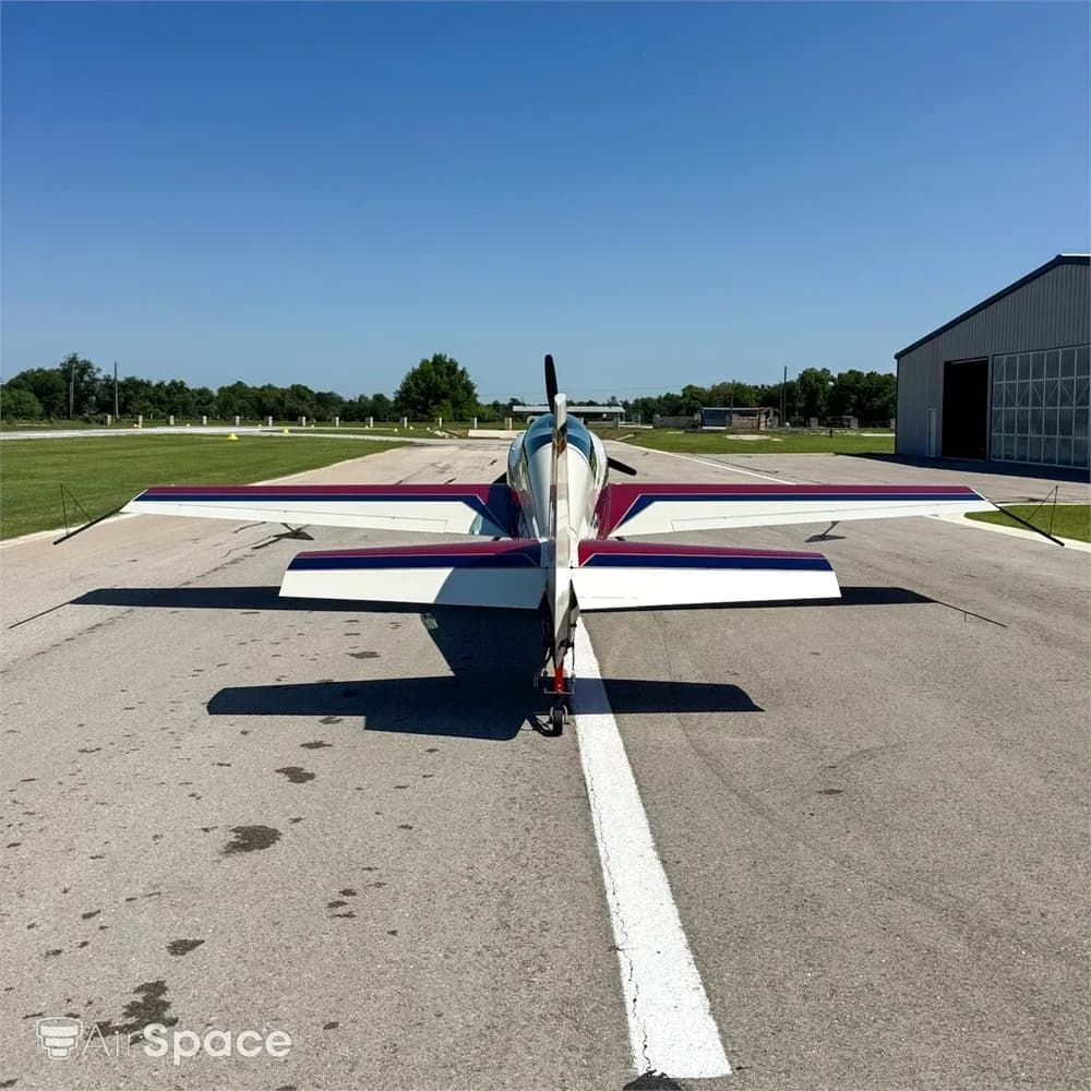 A direct rear view of the red, white, and blue Extra 300L aircraft, N251EC, showing its tail and wings on an airfield.