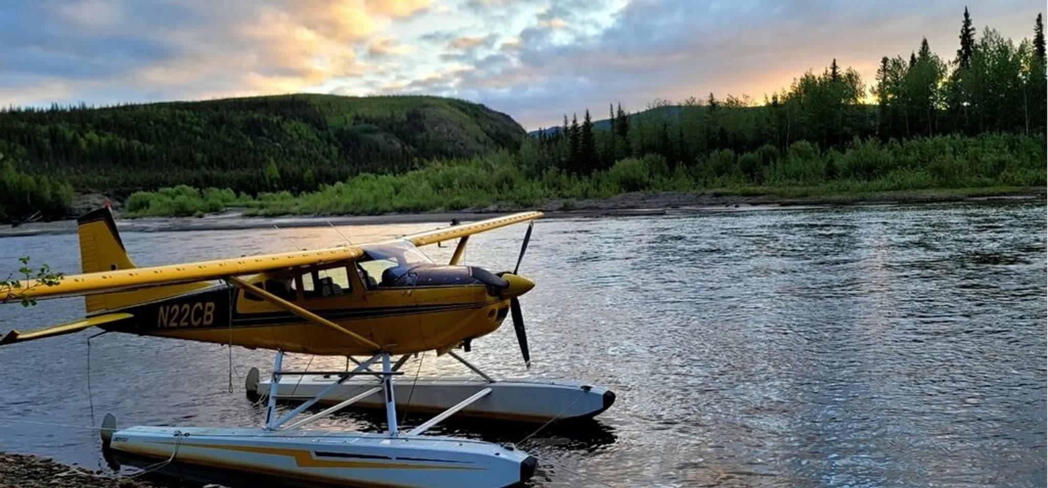 The yellow and brown floatplane N22CB is tied to a riverbank at sunset with forested hills in the background.