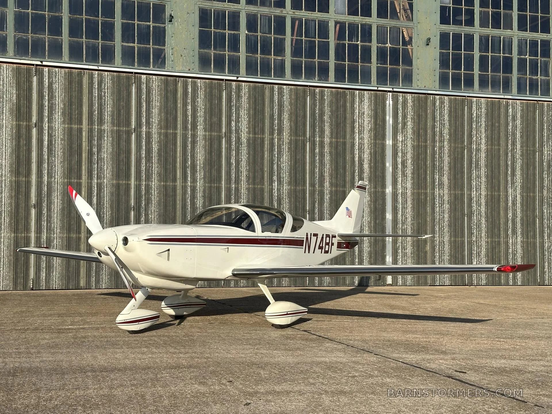 A white single-engine aircraft with a maroon stripe and registration N74BF is parked on concrete in front of a corrugated metal hangar.