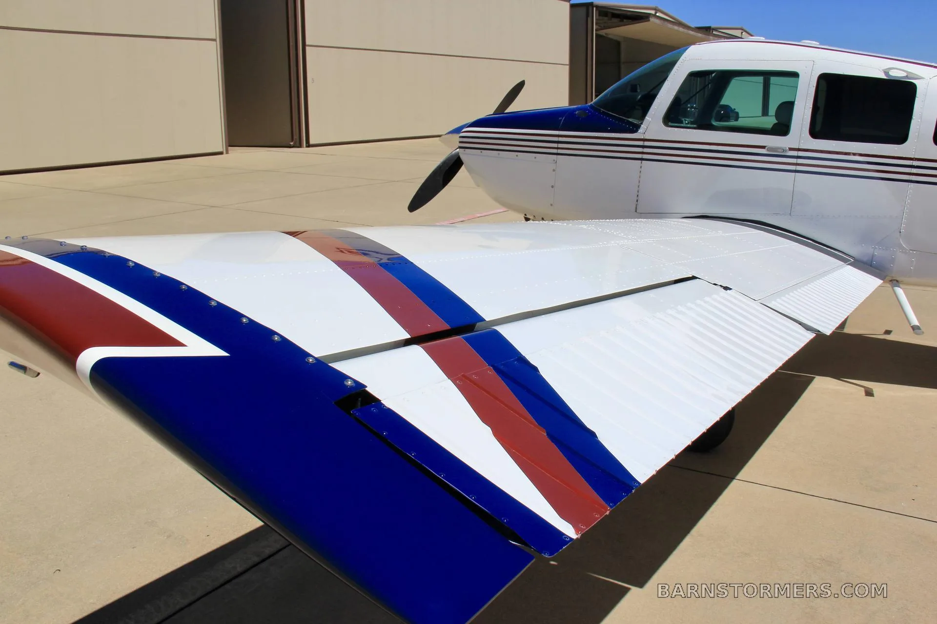 A close-up view of the wing and front fuselage of a white aircraft with blue and red stripes, showing the propeller and part of the cockpit.