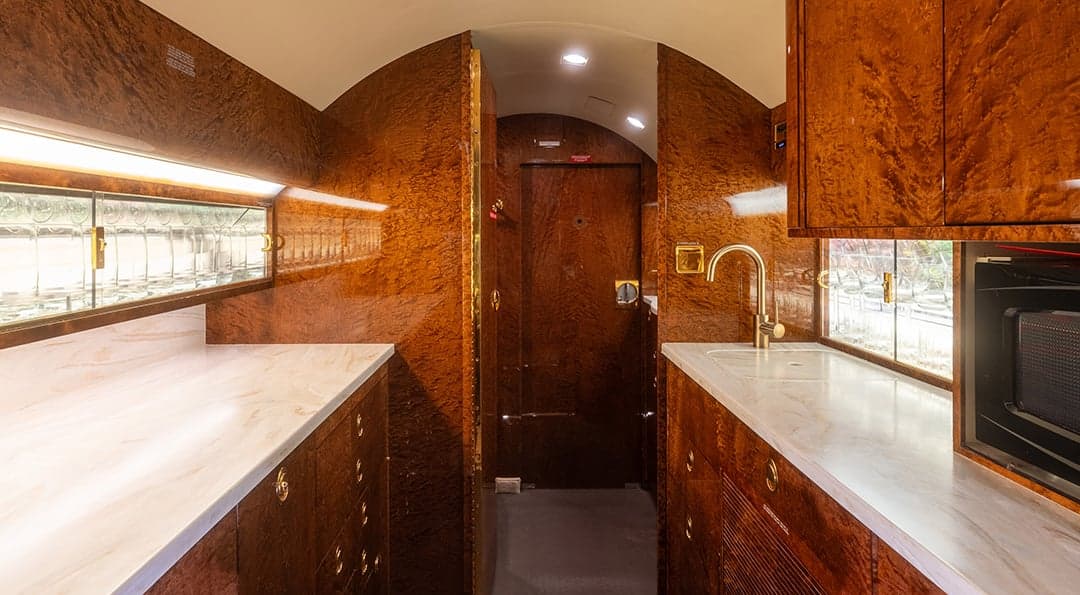 The galley of a private jet, featuring marble countertops, wood cabinetry, a golden faucet, and a microwave.