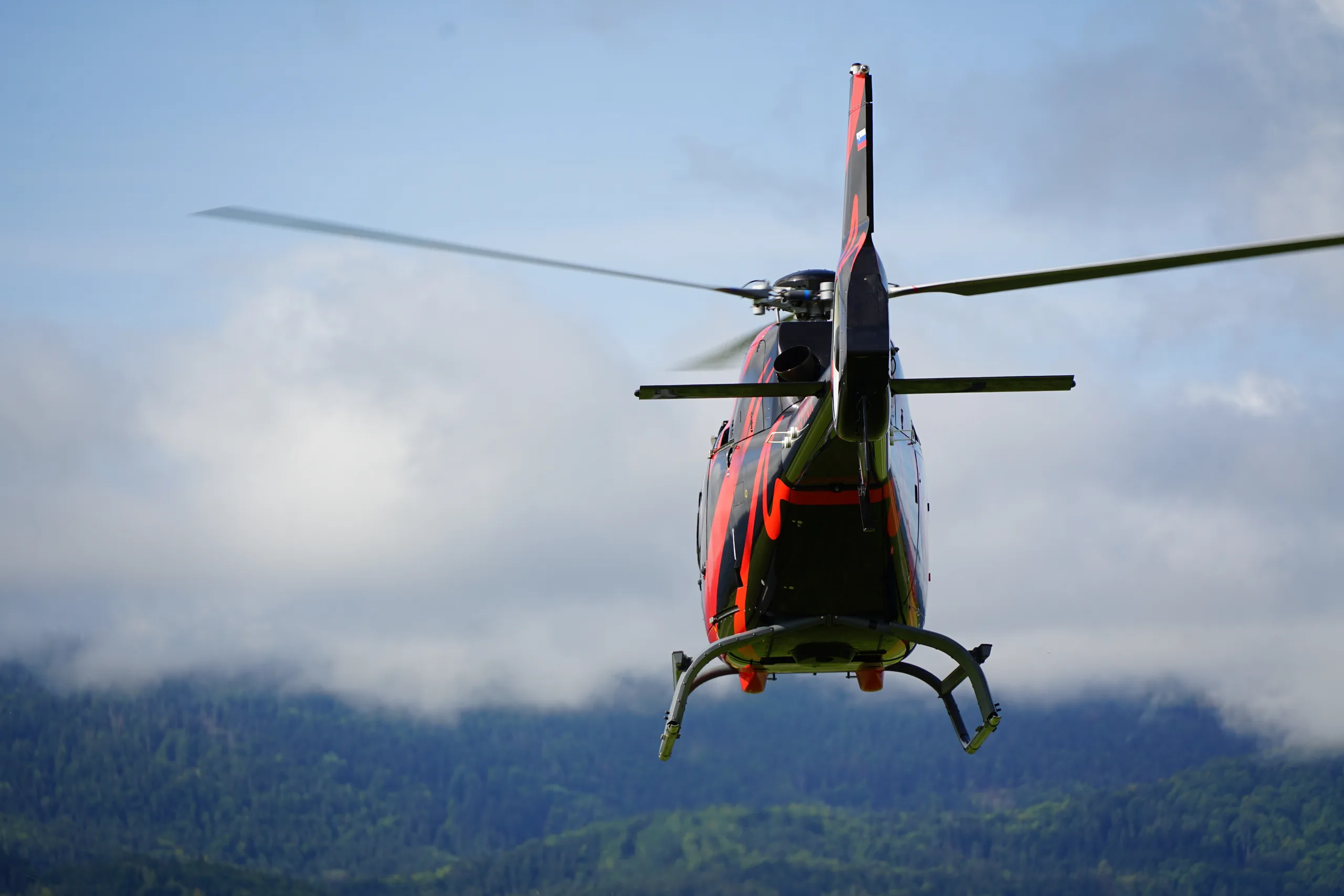 The rear of the black and red helicopter is shown in flight, with green hills in the background.