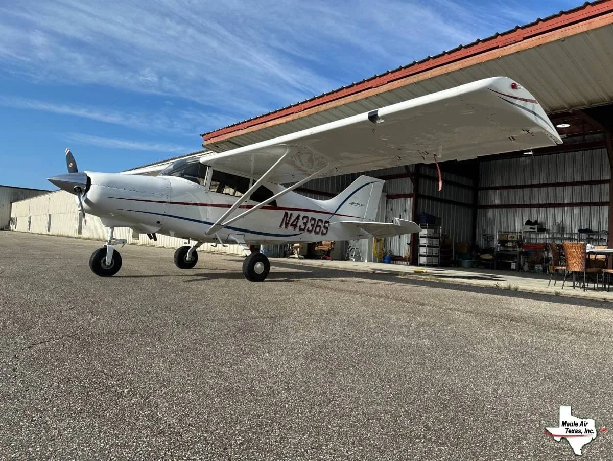 A white single-engine propeller aircraft with red and blue stripes, registration N4336S, parked on an asphalt tarmac next to an open hangar.