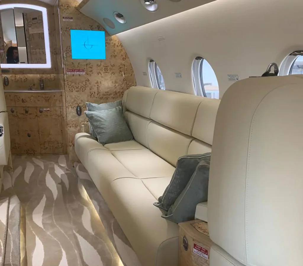 A perspective view of the private jet cabin showing a cream leather sofa, windows, and a patterned wall with a mirror.
