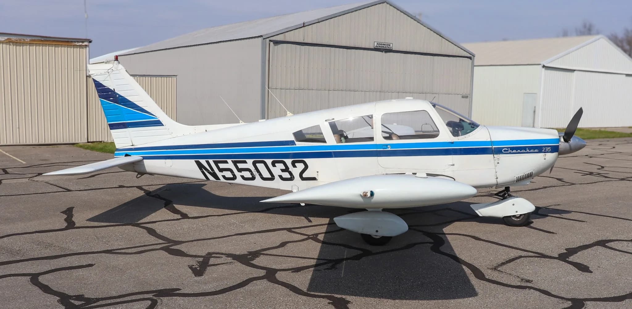A side view of the white Piper Cherokee 235 aircraft with blue stripes and registration N55032, parked on an asphalt ramp.