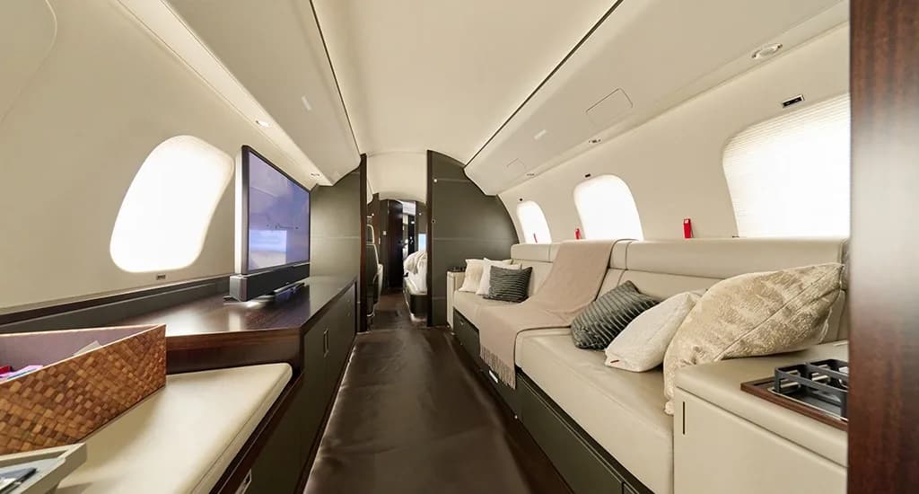 A private jet cabin featuring a long cream sofa, a large television screen, and dark wood cabinetry.