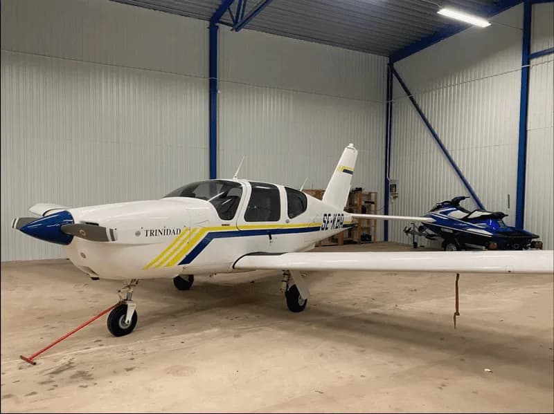 A white Socata TB-20 Trinidad aircraft, registration SE-KBA, with blue and yellow stripes, parked in a hangar.