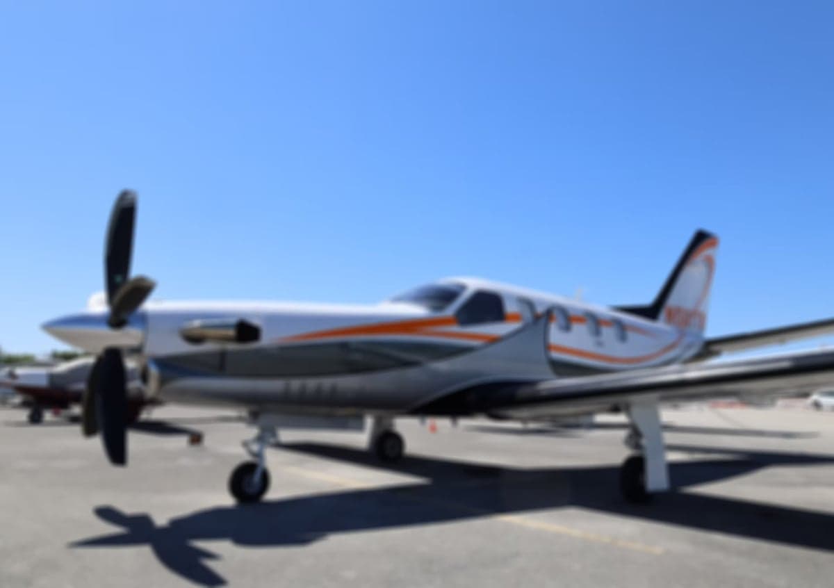 A blurry side view of a white and orange single-engine turboprop aircraft on an airfield under a clear blue sky.