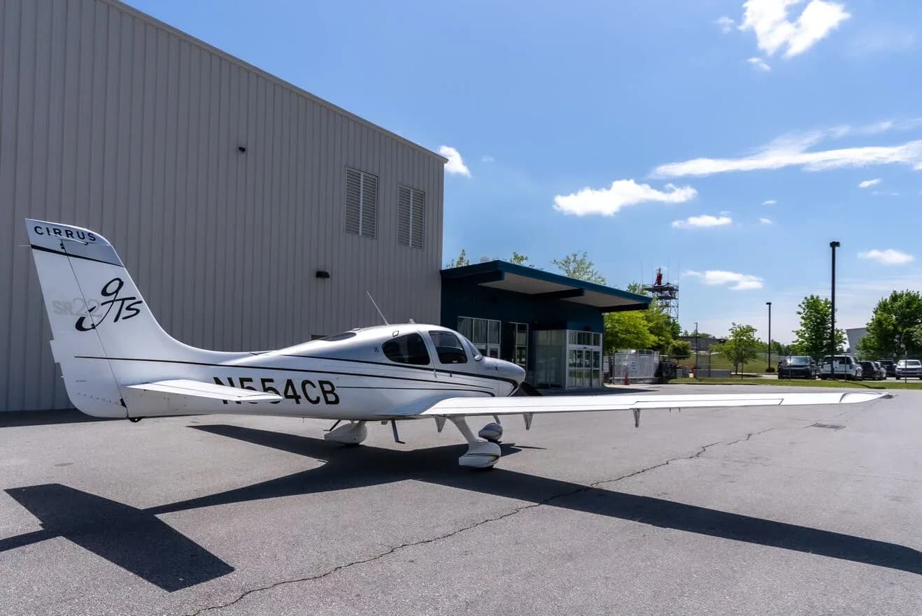 A rear three-quarter view of a silver and white Cirrus SR22T aircraft with registration N554CB parked next to a hangar.