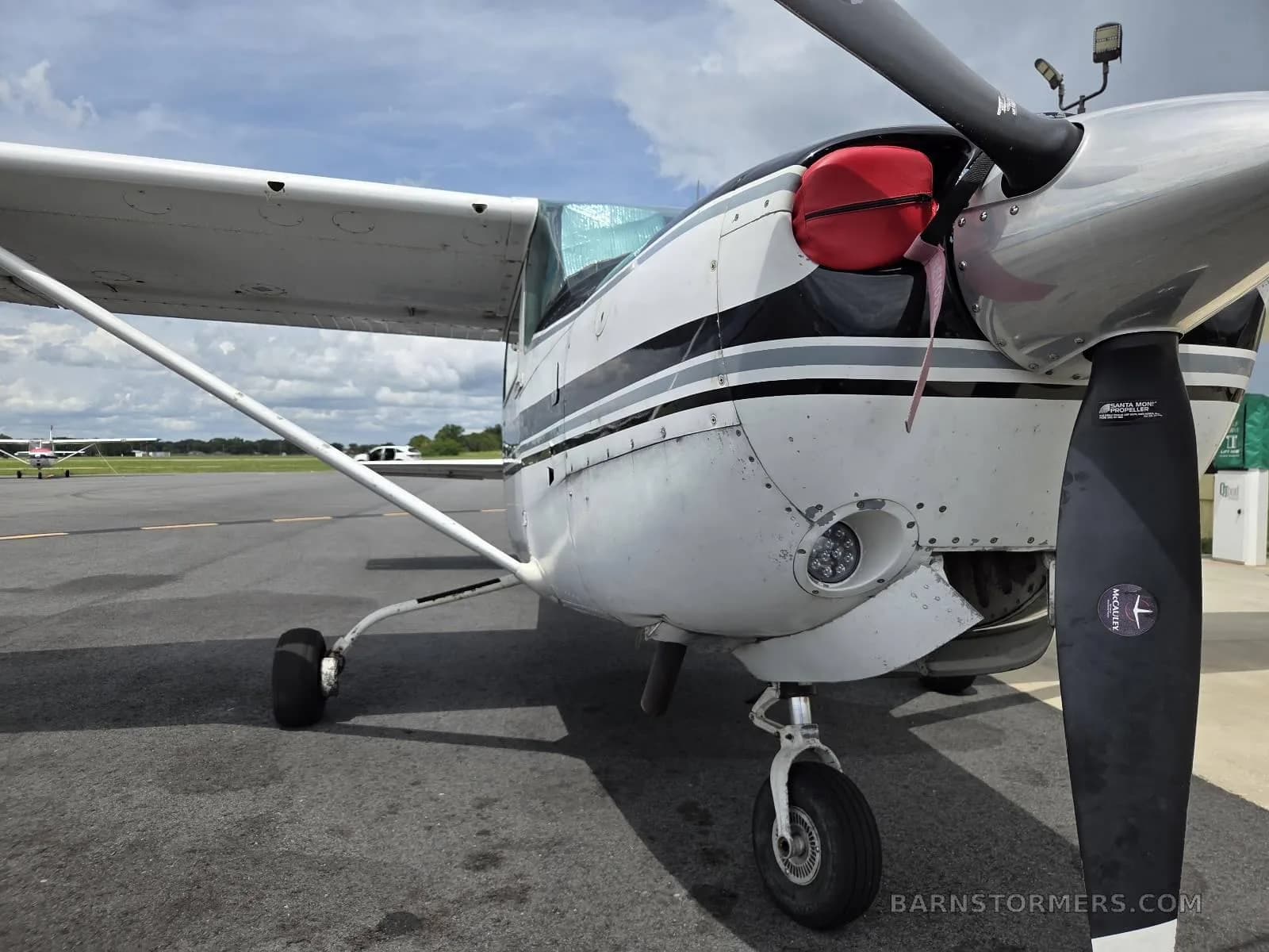 A close-up view of the front of a white and black Cessna aircraft, showing the propeller, nose cone with a red cover, and landing gear on an airfield.