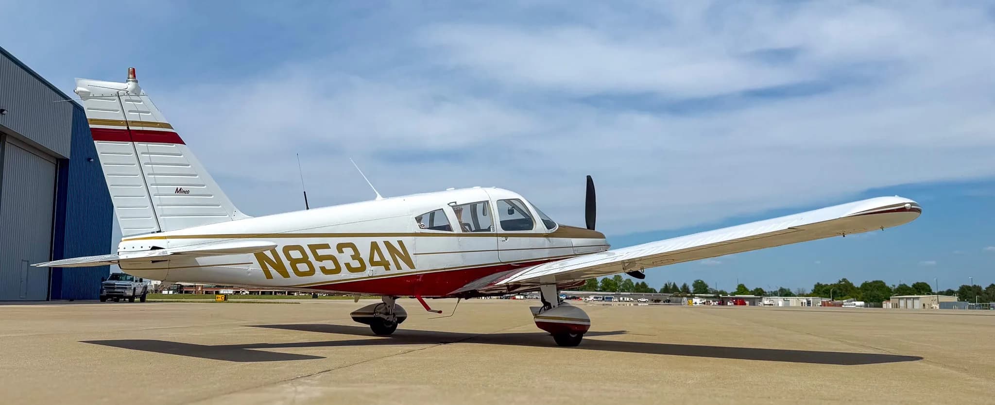 A side view of the white, red, and gold Piper Cherokee aircraft (N8534N) parked on a sunny airfield.