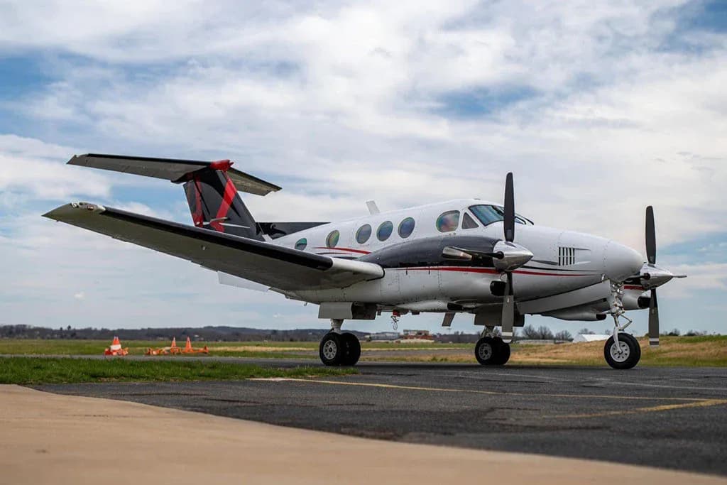 A white and black Beechcraft King Air with red accents on the tail, parked on an airport tarmac under a cloudy sky.