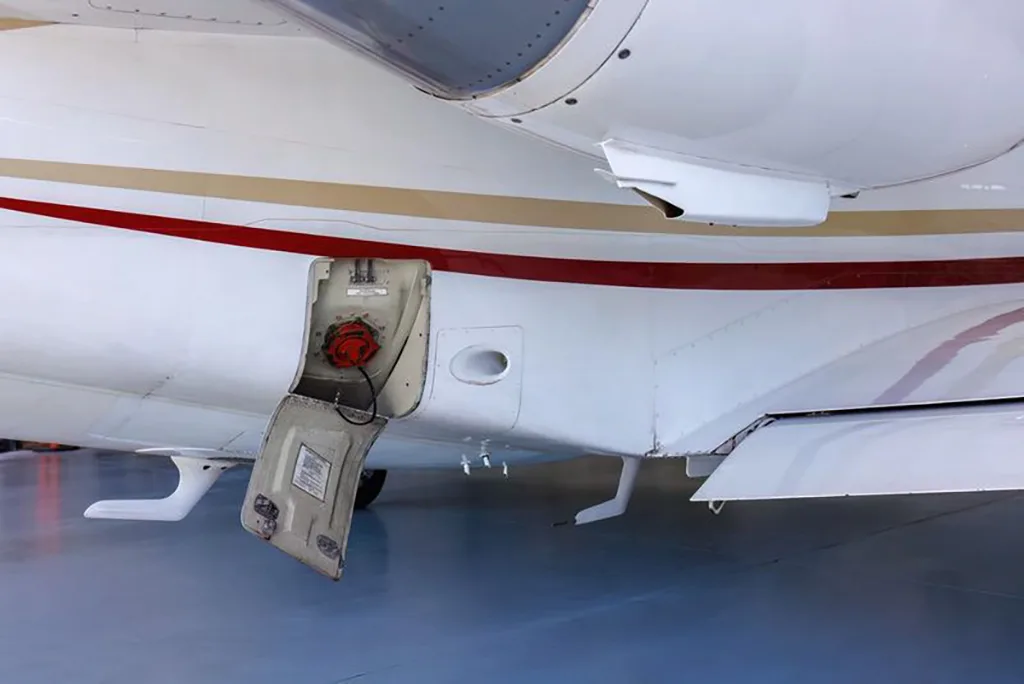 A close-up view of the white fuselage of a business jet, showing an open fuel port with a red cap.