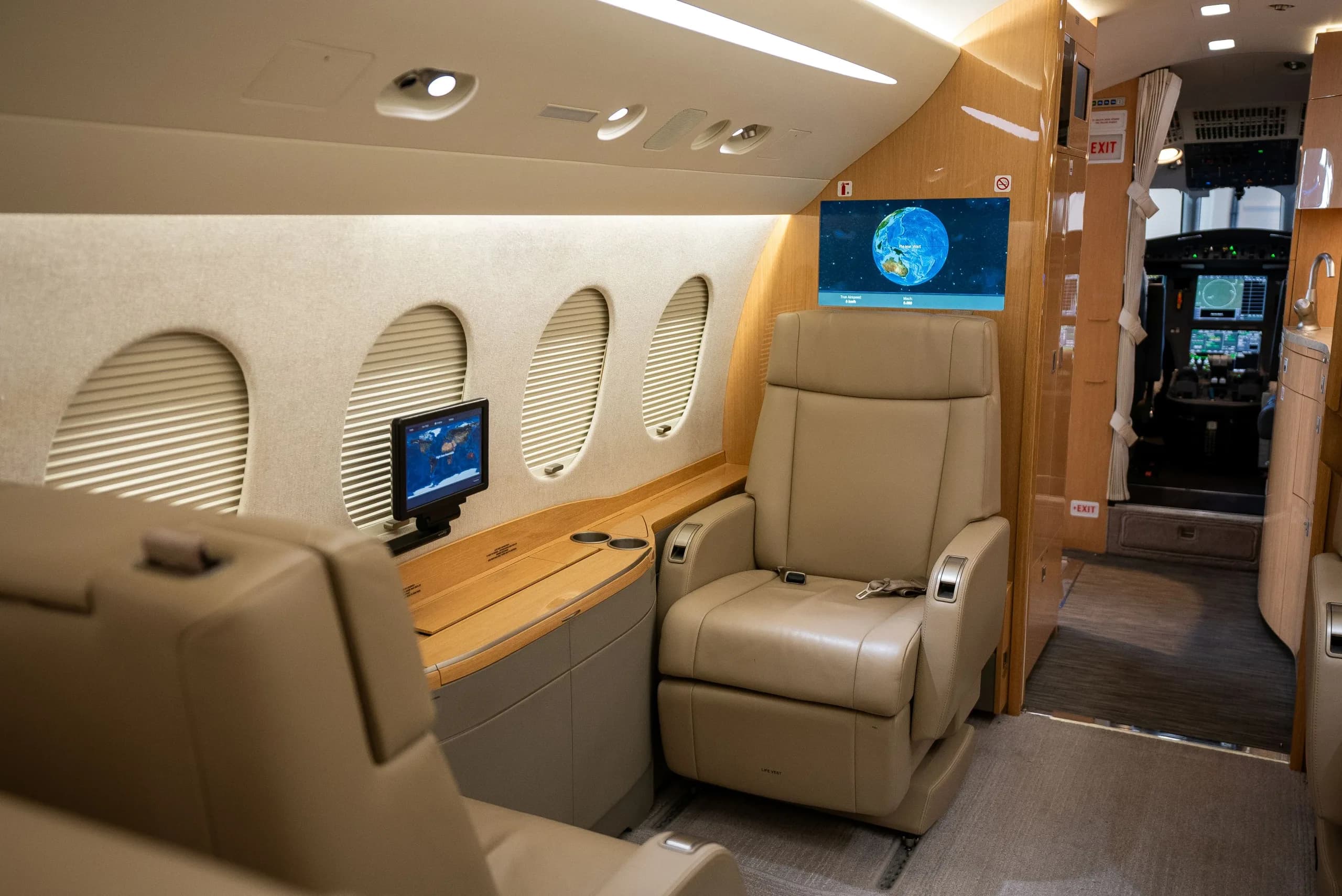 A view inside a private jet cabin with tan leather seats, wood trim, and a large screen displaying a map, looking towards the cockpit.