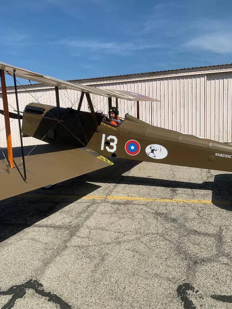 A side view of a brown biplane with a pilot in the cockpit, marked with "13" and a British roundel, parked on an asphalt apron.