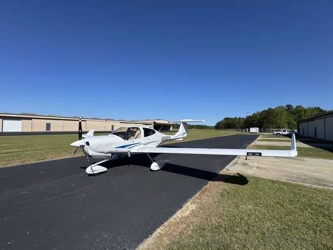 A white Diamond DA40 aircraft with blue stripes is parked on an airport ramp under a clear blue sky.