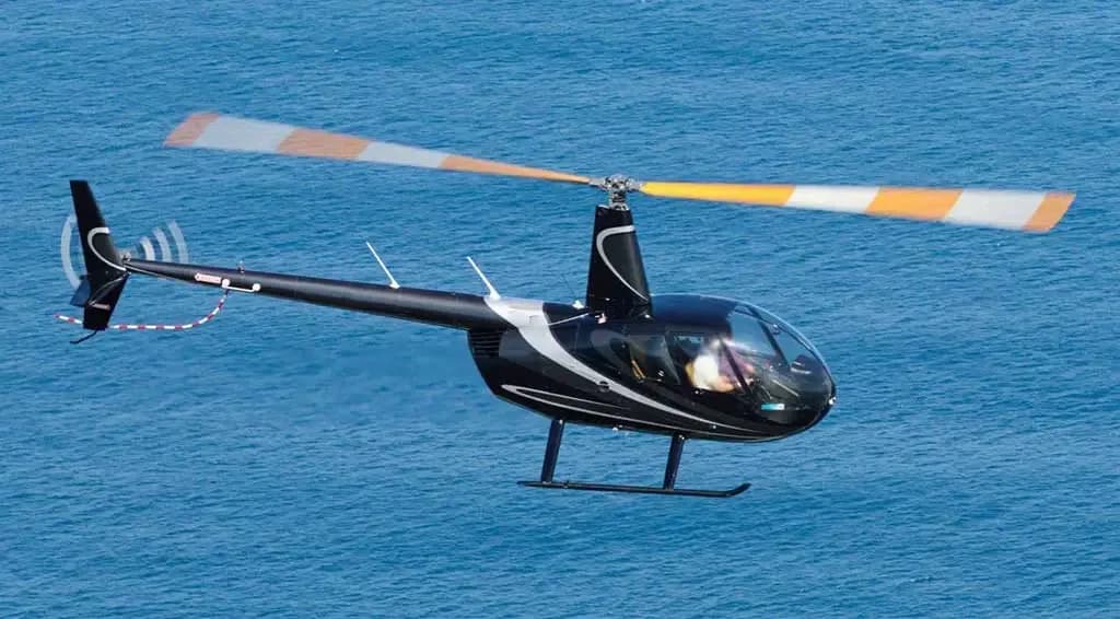 A black Robinson R44 helicopter with silver stripes flying over blue water.