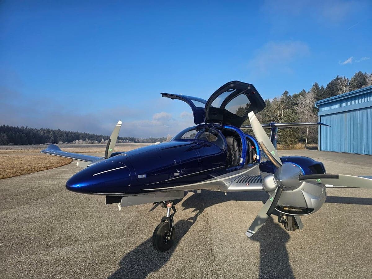 A blue and silver Diamond DA62 aircraft is parked on the tarmac with its gull-wing doors open.