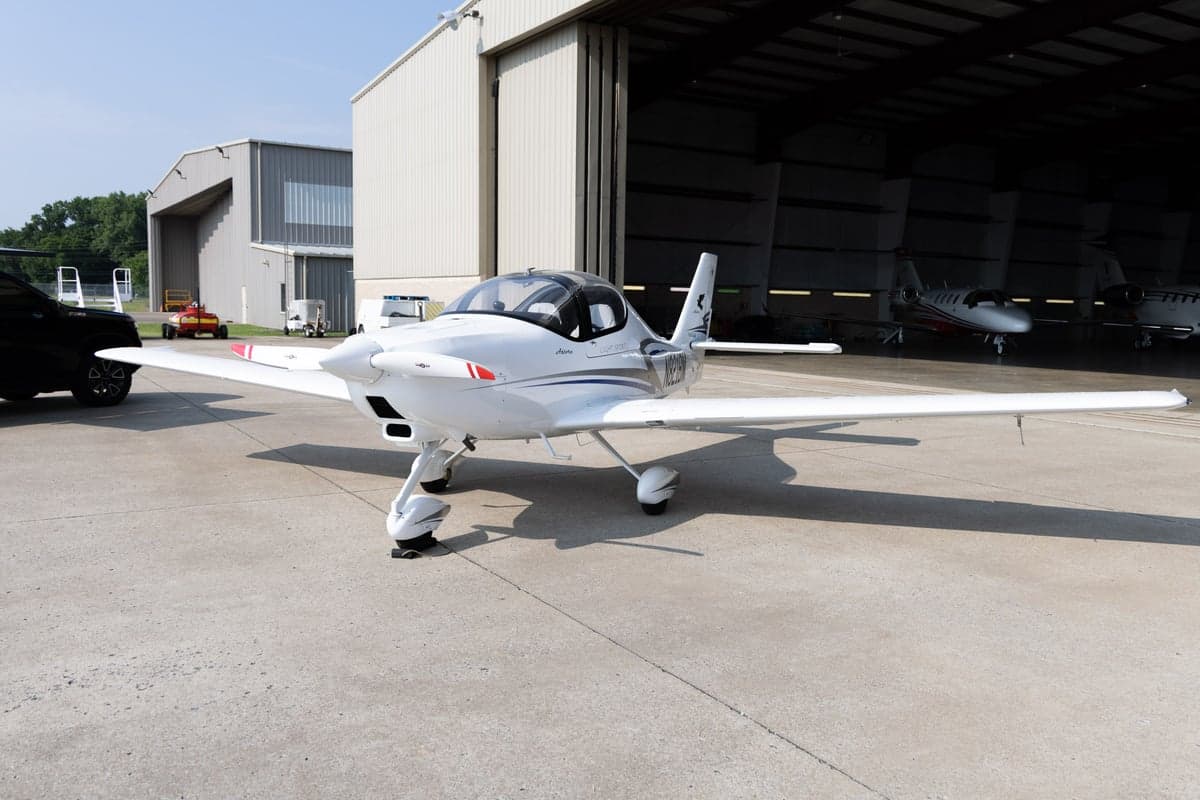 A white Tecnam Astore light sport aircraft with blue and grey stripes parked on a tarmac next to a large hangar.