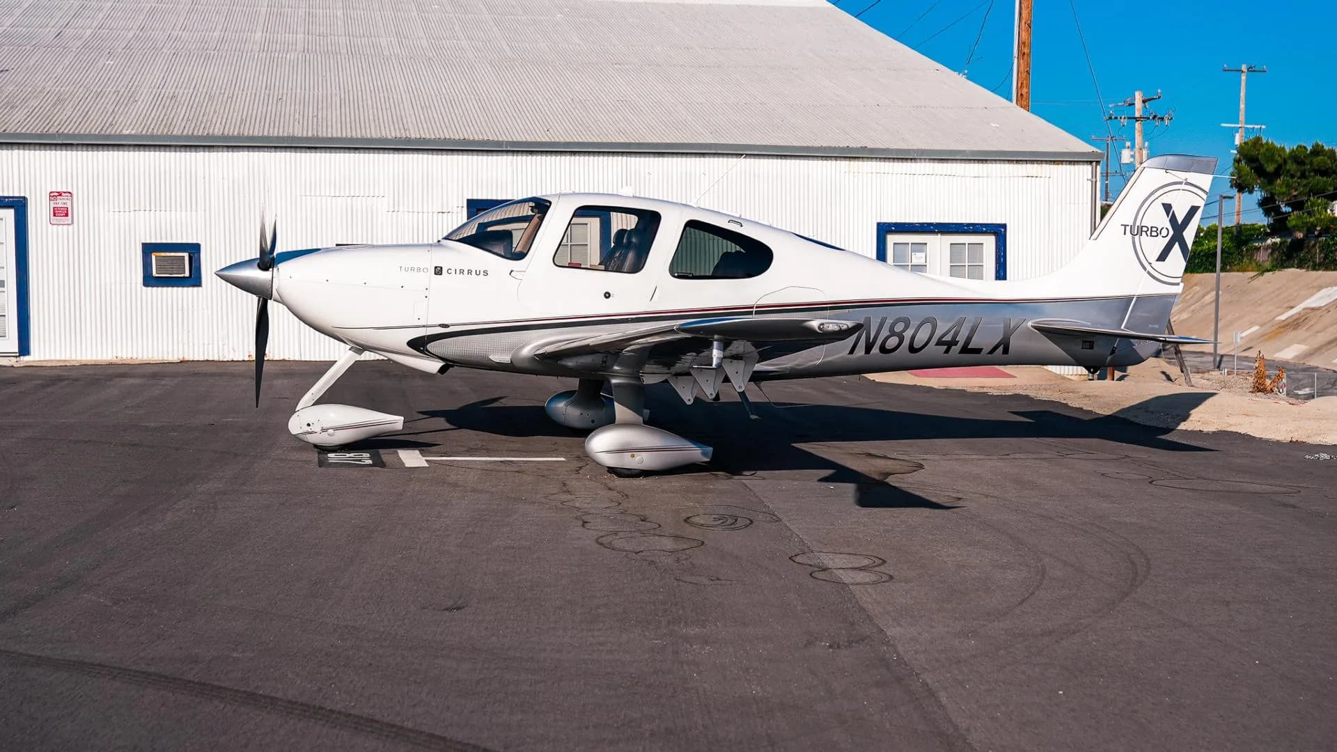 A side view of the white and silver Cirrus SR22T aircraft N804LX parked in front of a white hangar.