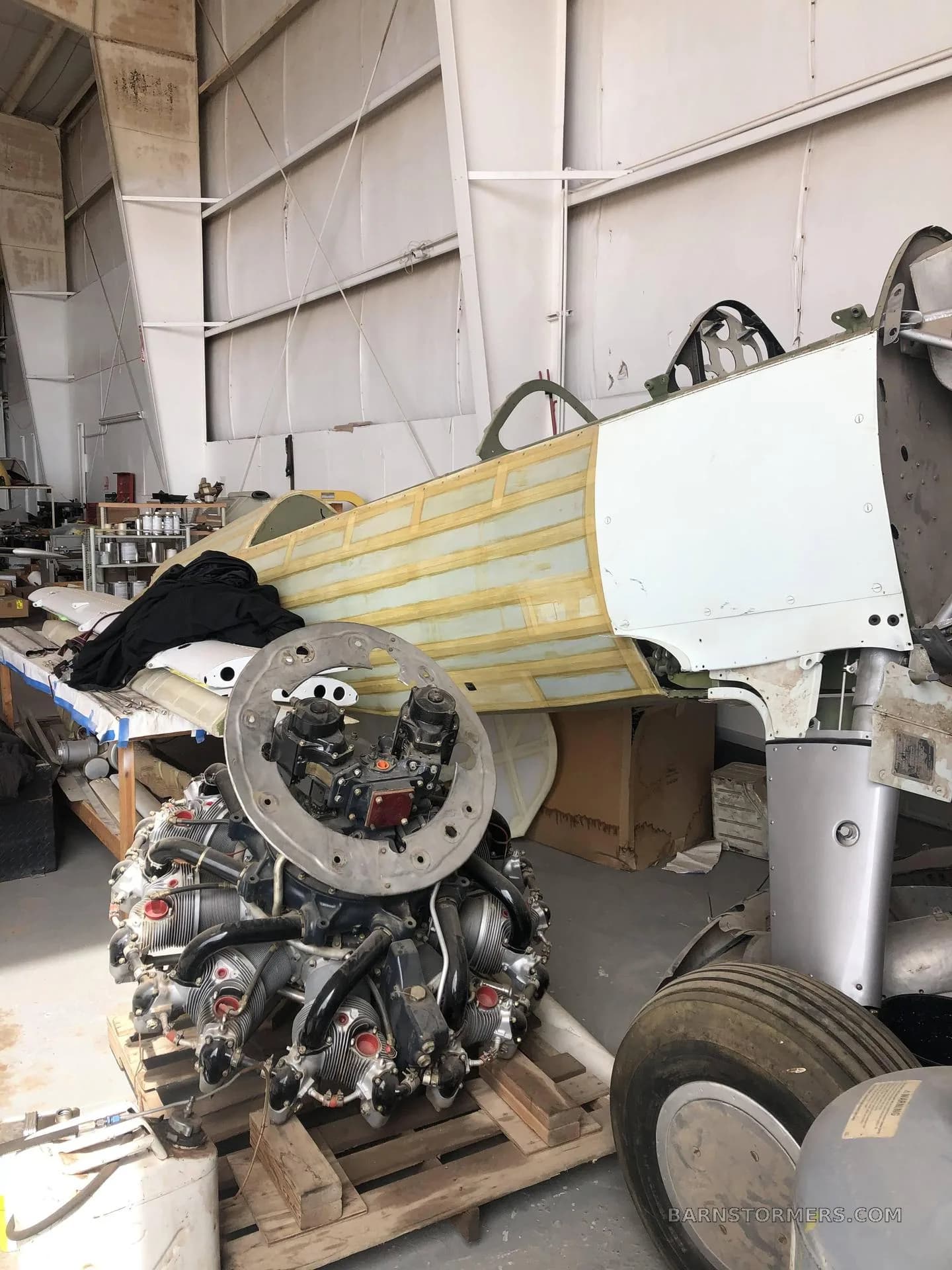 A radial aircraft engine on a pallet in the foreground, with a partially stripped fuselage section in the background inside a hangar.