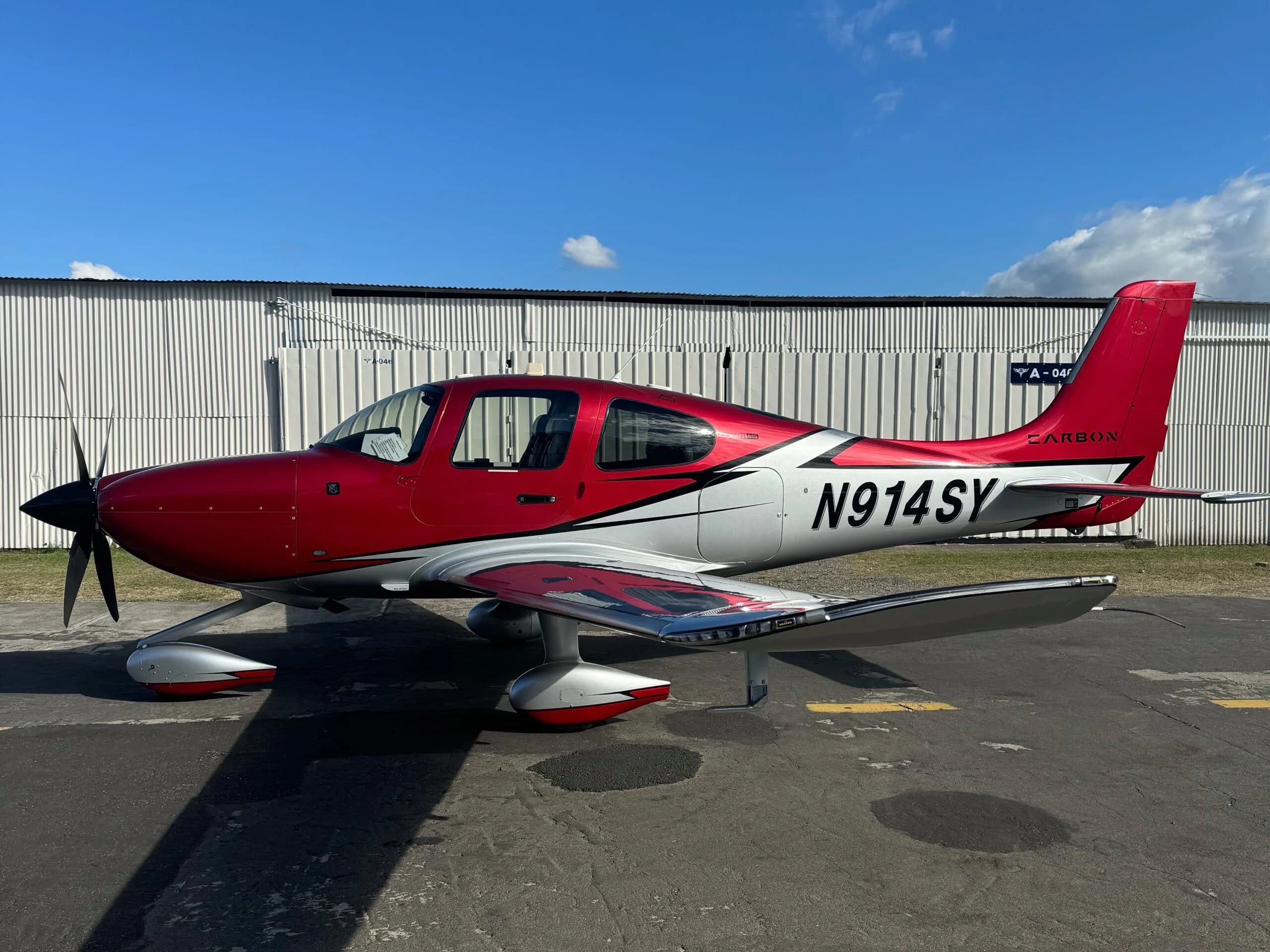 A red and silver Cirrus SR22 aircraft with registration N914SY parked on an asphalt apron in front of a white hangar.