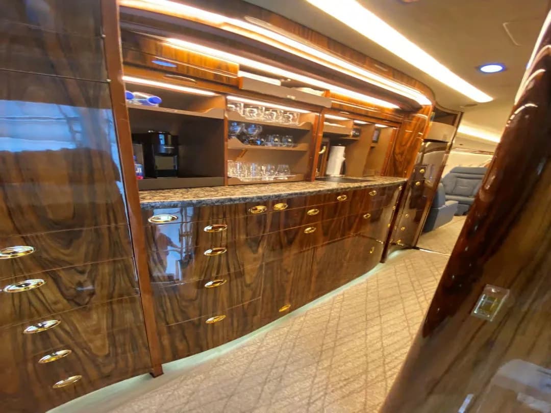 A luxurious aircraft galley features dark wood cabinetry with gold hardware, a granite countertop, and glassware.