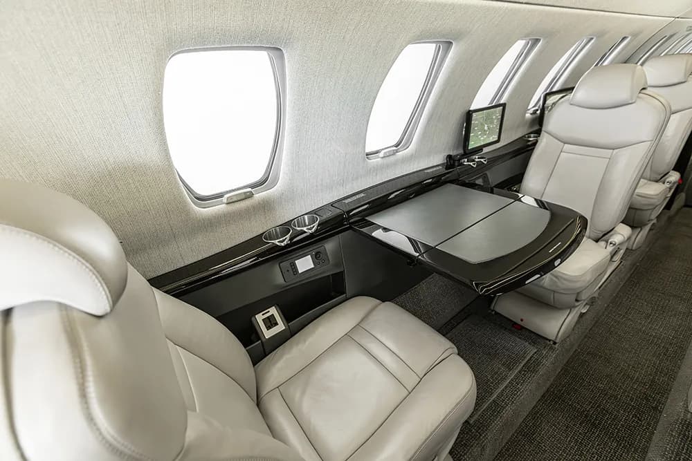 A close-up of the private jet's cabin interior, highlighting a fold-out table and light grey leather seats next to windows.