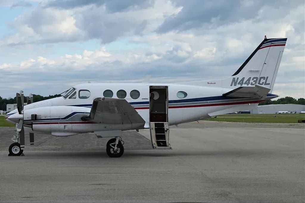 A white Beechcraft King Air C90GTx with blue and red stripes, parked on the tarmac with its passenger door open.