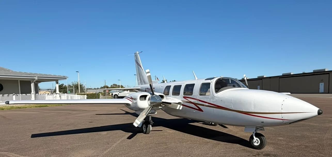 A front-quarter view of a white twin-engine propeller aircraft with red and gold stripes, registration N701KE, parked on an airfield.