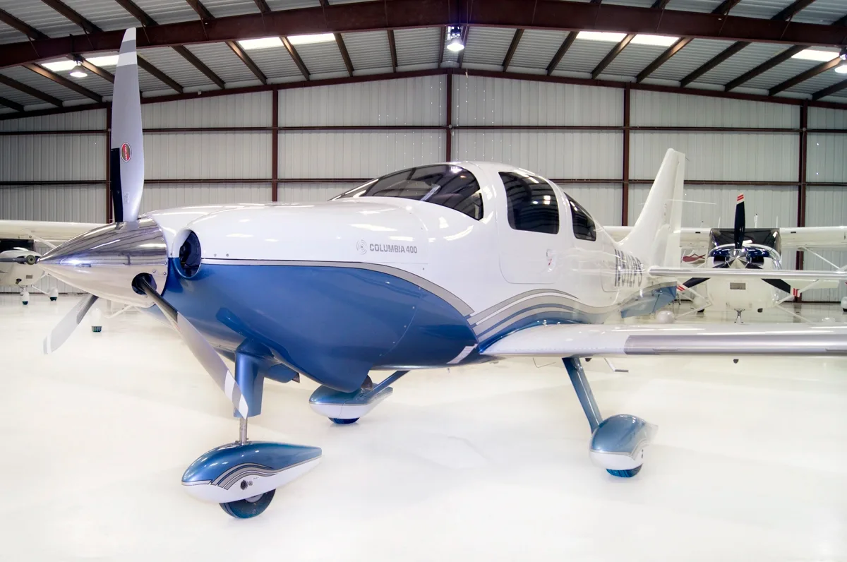 A white and blue Columbia 400 aircraft is parked in a hangar, viewed from the front-left.