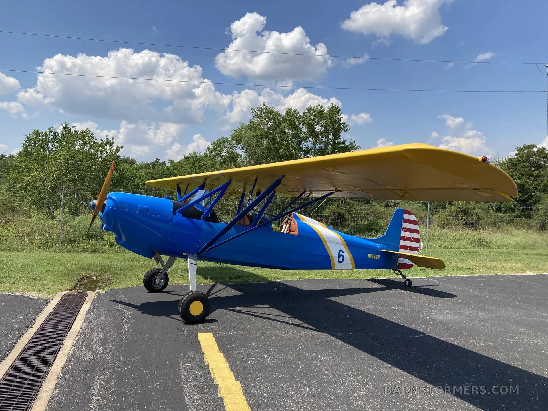 A blue and yellow biplane with '6' on the side, parked on an asphalt runway under a blue sky with clouds.