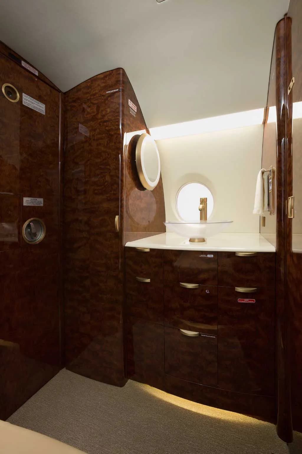 The private jet's lavatory area, featuring a vanity with a sink, mirror, and polished wood cabinetry.